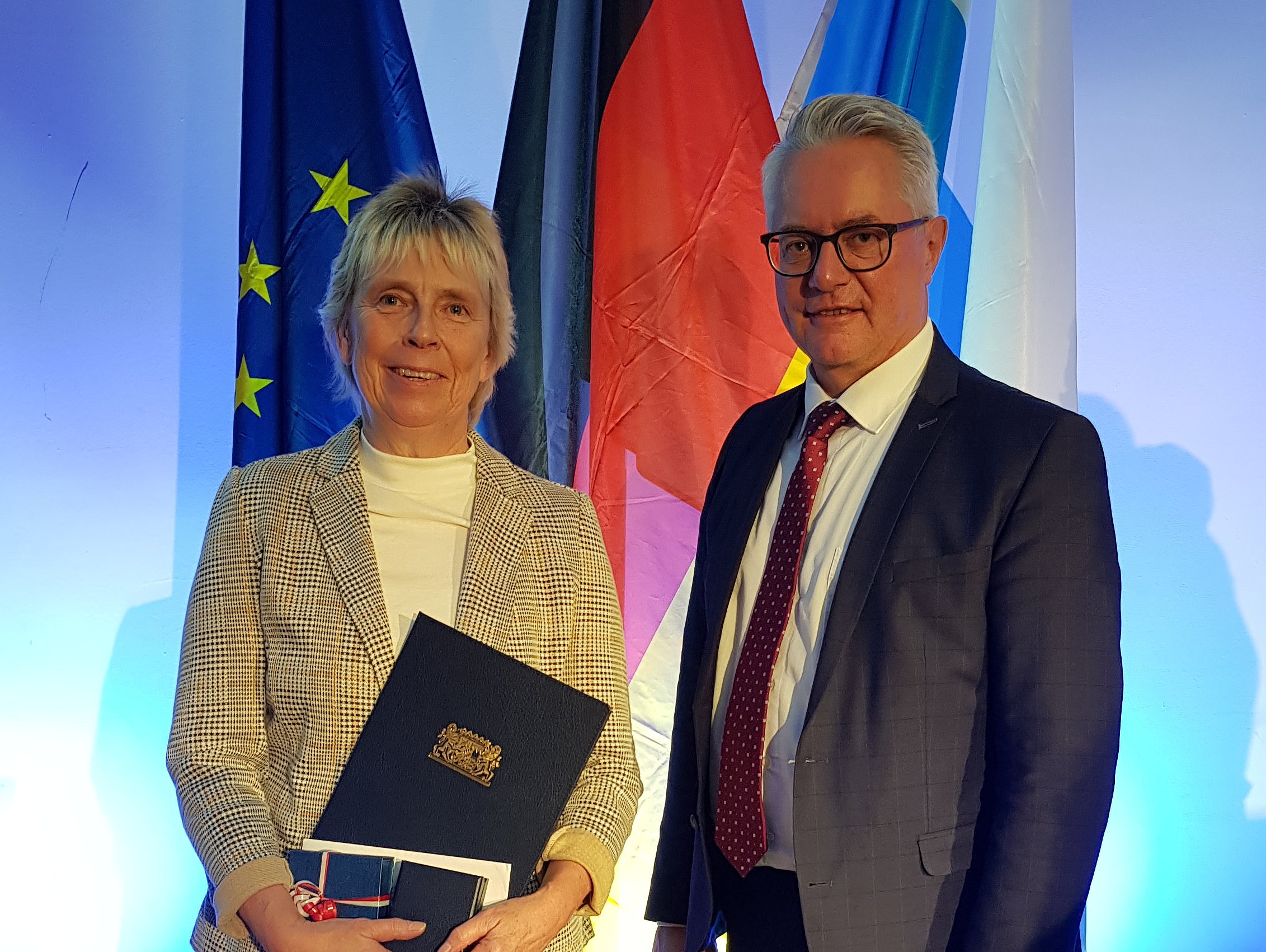 Uta Reif mit Bürgermeister Stefan Pöhlmann während der Feierstunde in Nürnberg Vor einem blauen Hintergrund stehen drei Fahnen, die europäische, die deutsche sowie die bayerische Fahne. Wiederum davor stehen eine Frau, die mit dem Ehrenamtspreis ausgezeichnet wurde, und der Helmbrechtser Bürgermeister.