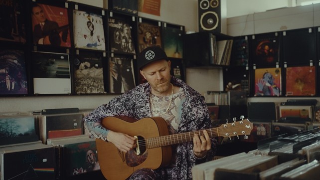 Angela Aux Der Musiker Angela Aux steht mit seinem Blick auf seine Gitarre gerichtet in einem Tonstudio, hinter ihm überall Schallplatten aufgereiht