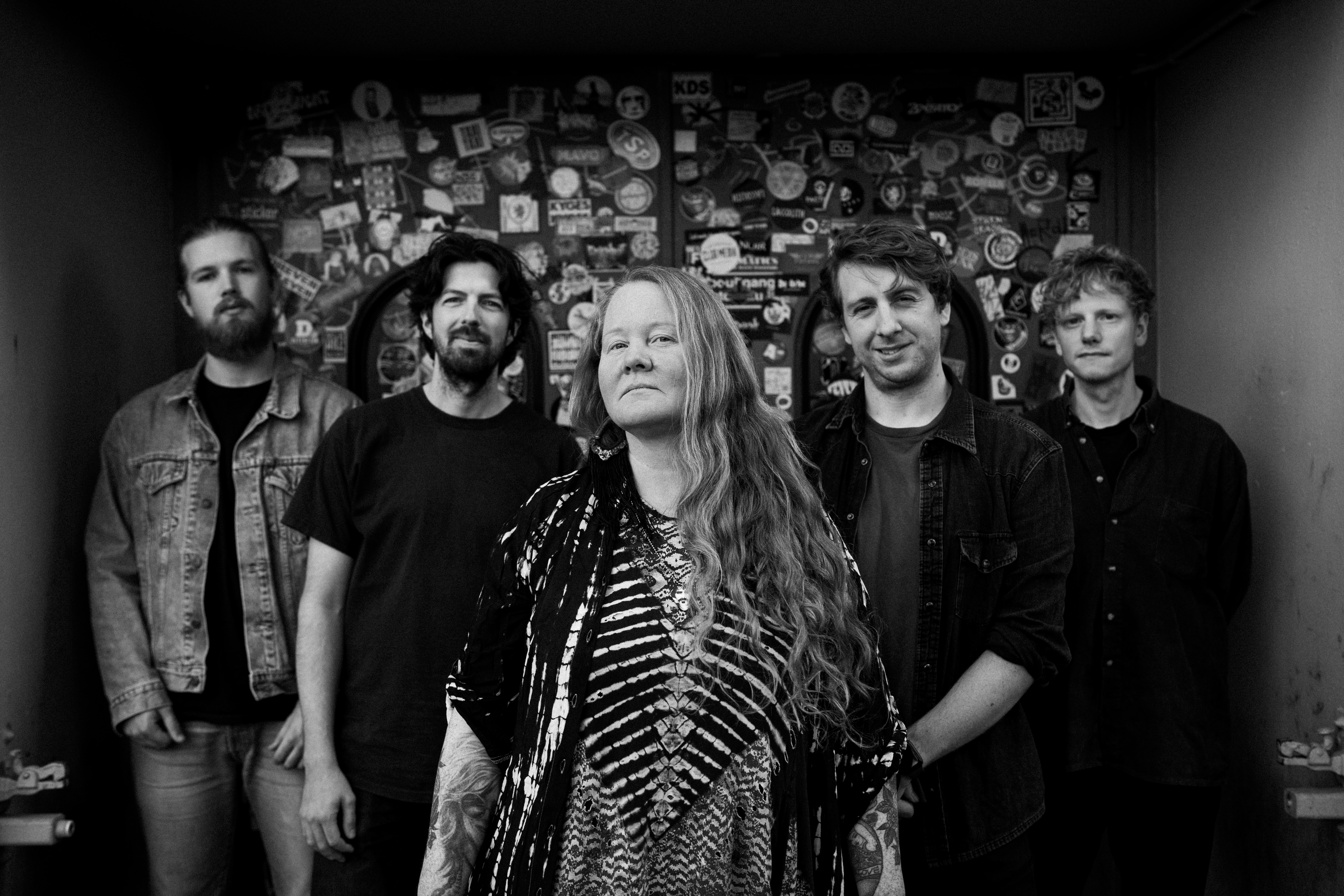 Layla Zoe Schwarz-Weiß Foto der fünfköpfigen Band Layla Zoe vor einer Wand mit zahllosen Stickern und Aufklebern